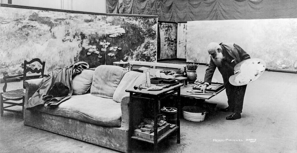 モノクロ写真：モネがアトリエでパレットを持って筆を選んでいるところ。睡蓮の作品に囲まれている
